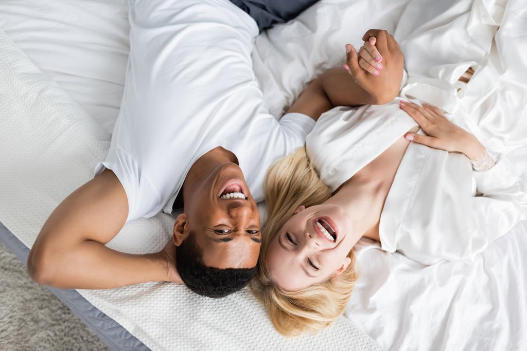 top view of excited multiethnic couple holding hands and laughing at camera on white bedding - Photo, Image