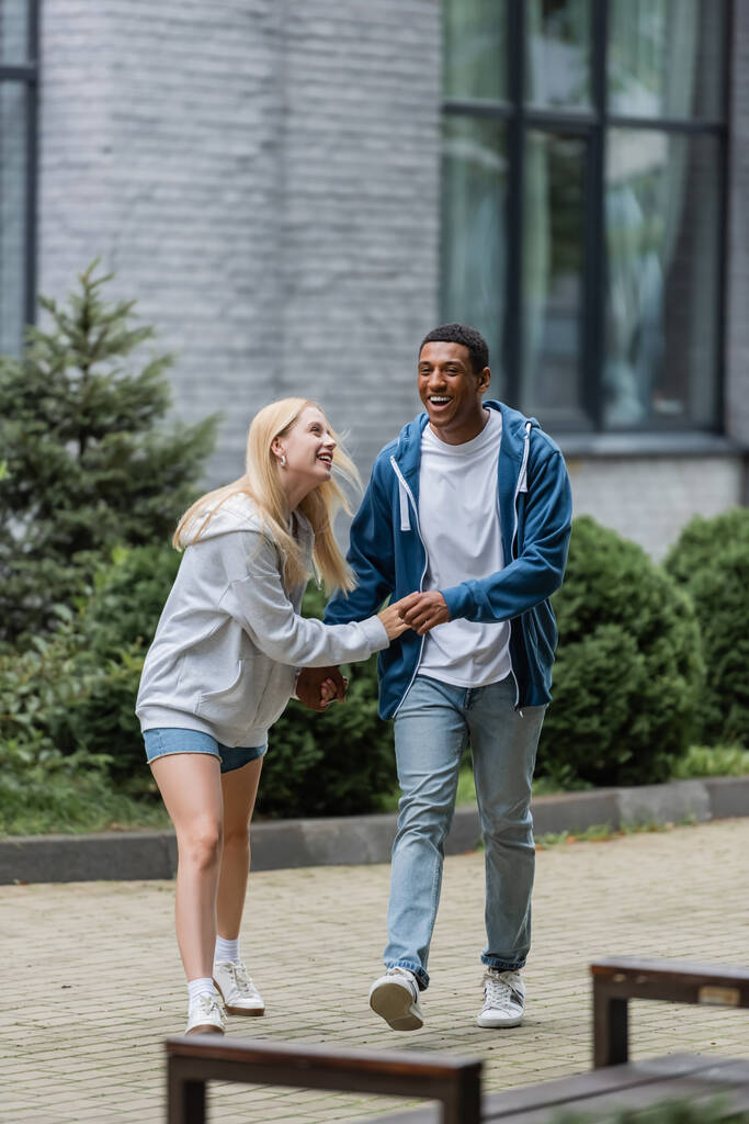 longitud completa de alegre pareja interracial caminando y riendo en la calle urbana - Foto, Imagen