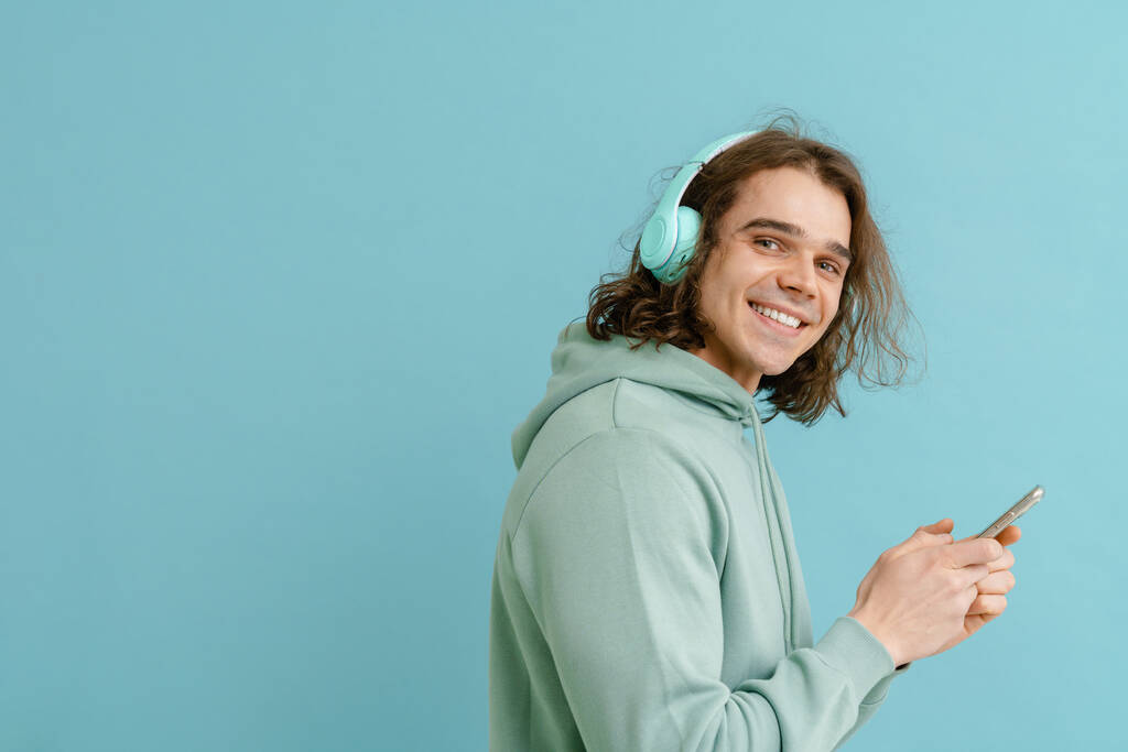 Jeune homme souriant aux cheveux longs dans les écouteurs tenant le téléphone et regardant à la caméra sur fond bleu isolé - Photo, image