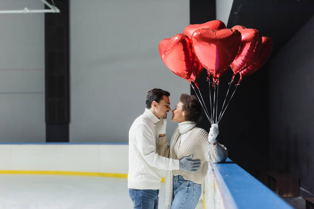 Side näkymä iloinen afrikkalainen amerikkalainen nainen suudella poikaystävä ja tilalla sydämen muotoinen ilmapalloja jäähalli  - Valokuva, kuva