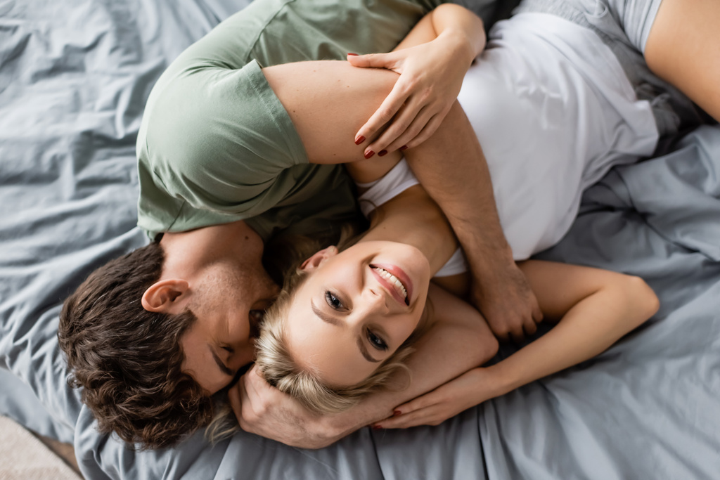 Top pohled na veselý muž objímání přítelkyně v pyžamu při pohledu na kameru na posteli  - Fotografie, Obrázek