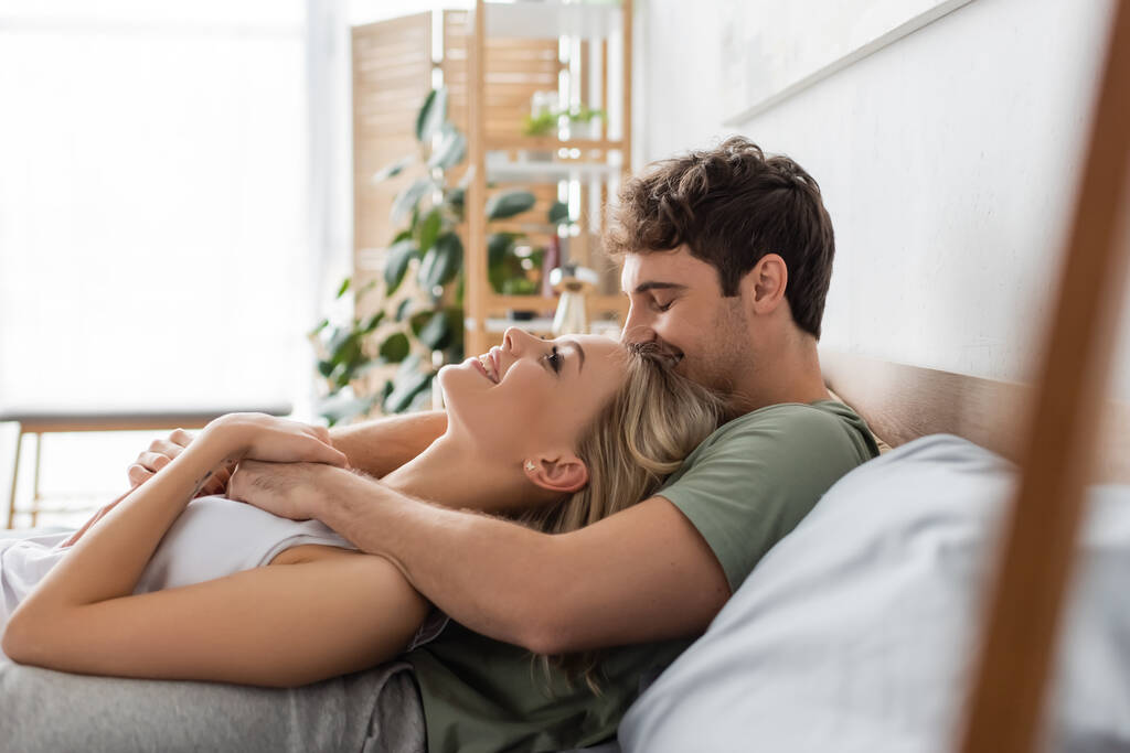 Vista lateral de un hombre joven besando a su novia rubia en la cama por la mañana  - Foto, imagen