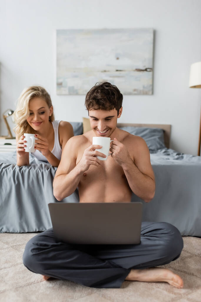 Positivo hombre musculoso sosteniendo la taza de café y mirando a la computadora portátil cerca de la novia en la cama por la mañana  - Foto, imagen