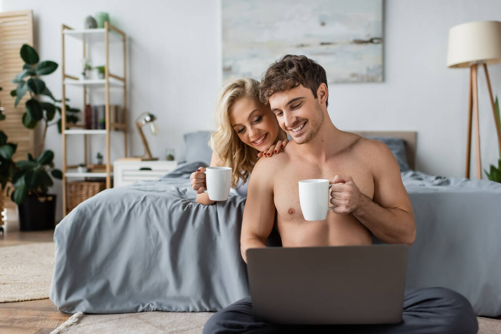 Coppia sorridente con tazze di caffè guardando il computer portatile in camera da letto al mattino  - Foto, immagini