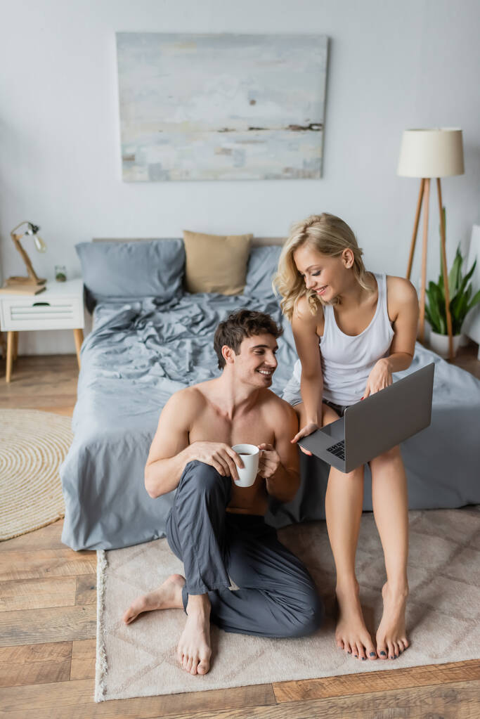 Uśmiechnięta kobieta w piżamie trzyma laptopa blisko seksownego chłopaka z kawą w sypialni  - Zdjęcie, obraz