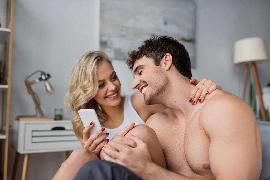 Uśmiechnięta blondynka trzyma smartfona i przytula chłopaka bez koszulki w sypialni  - Zdjęcie, obraz