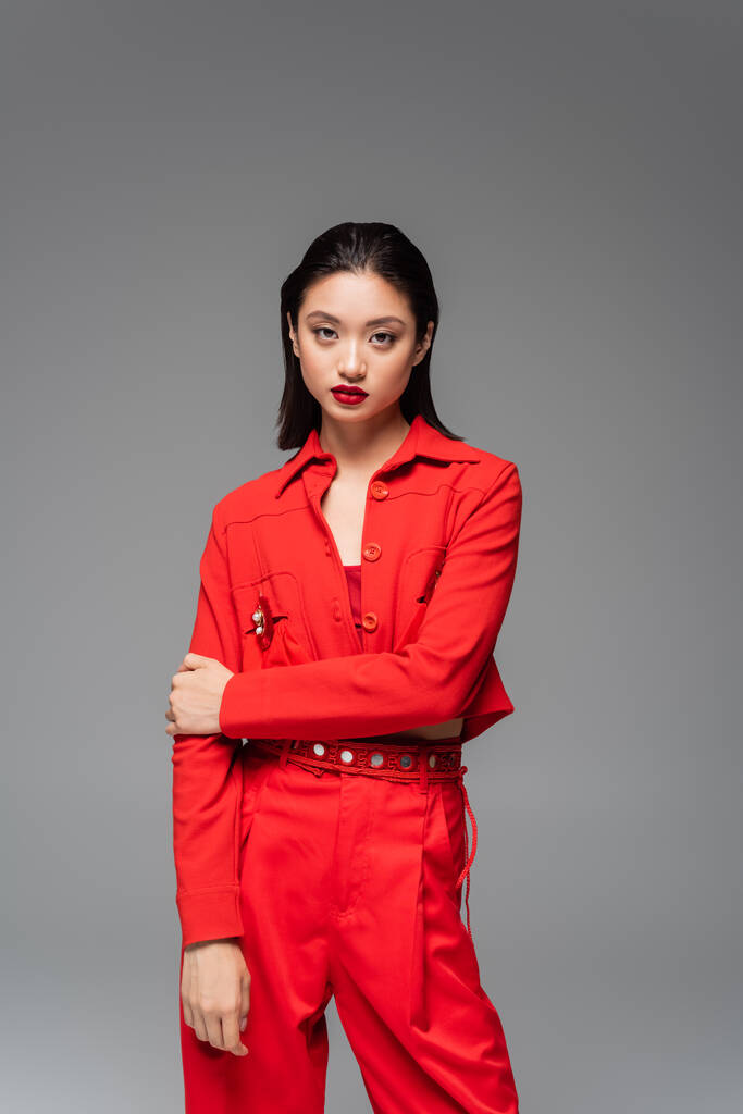 brunette asian woman in red trendy suit looking at camera isolated on grey - Photo, Image
