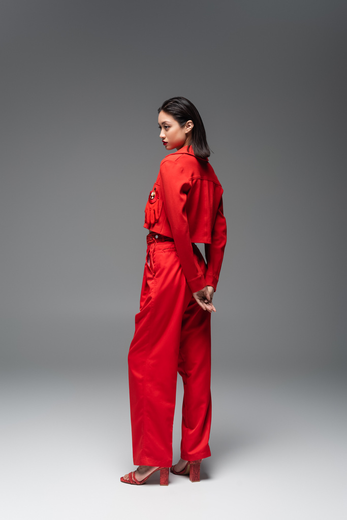 πλήρες μήκος της ασιατικής γυναίκας σε κόκκινο σακάκι και παντελόνι στέκεται σε γκρι φόντο - Φωτογραφία, εικόνα