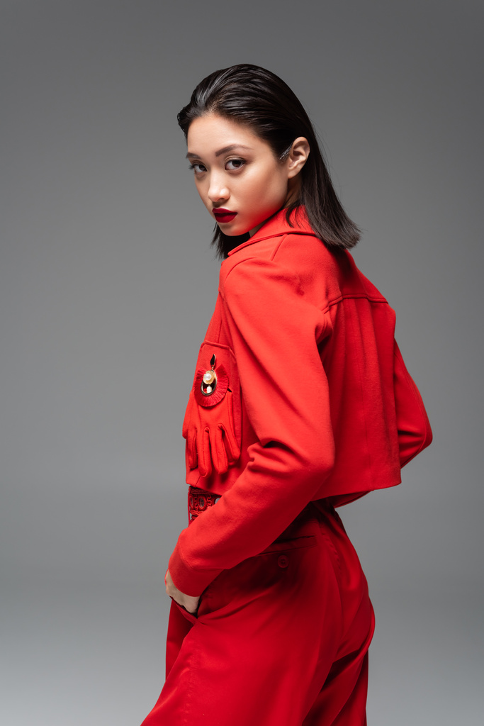 fashionable asian woman in red attire holding hand in pocket and looking at camera isolated on grey - Photo, Image