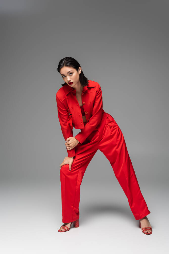 volle Länge der stilvollen asiatischen Frau in roter Jacke und Hose, die in die Kamera schaut, während sie auf grauem Hintergrund posiert - Foto, Bild