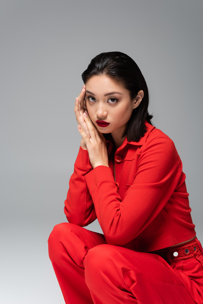 stylish asian woman in red elegant attire sitting on haunches with hands near face isolated on grey - Photo, Image