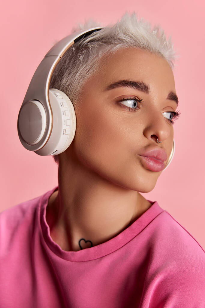 ヘッドフォンで音楽を聴いている短い髪と灰色の目を持つ若いスタイリッシュな女性は、ピンクの背景に隔離されたポーズ。若者、美しさ、ファッション、ライフスタイル、感情、表情の概念。広告 - 写真・画像