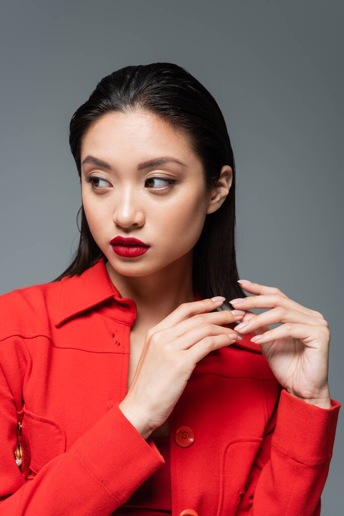 красивая азиатская женщина с макияжем и красными губами в модной куртке и глядя в сторону изолированы на сером - Фото, изображение