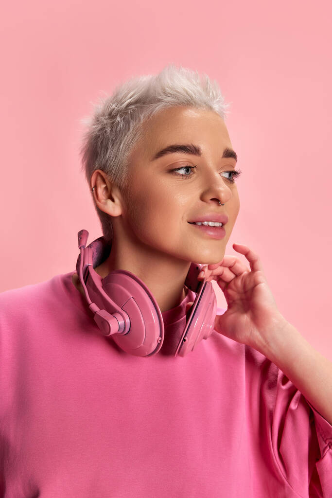 Porträt einer jungen, stylischen Frau mit kurzen Haaren, die mit Kopfhörern vor rosa Hintergrund posiert. Jugendlicher Lebensstil. Konzept der Jugend, Stil, Schönheit, Mode, Musik, Emotionen, Mimik. Anzeige - Foto, Bild