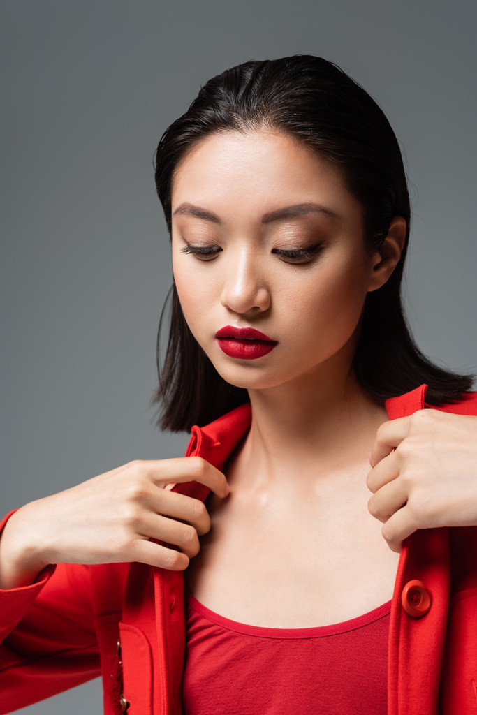 joven asiático mujer con rojo labios tocando elegante chaqueta aislado en gris - Foto, Imagen