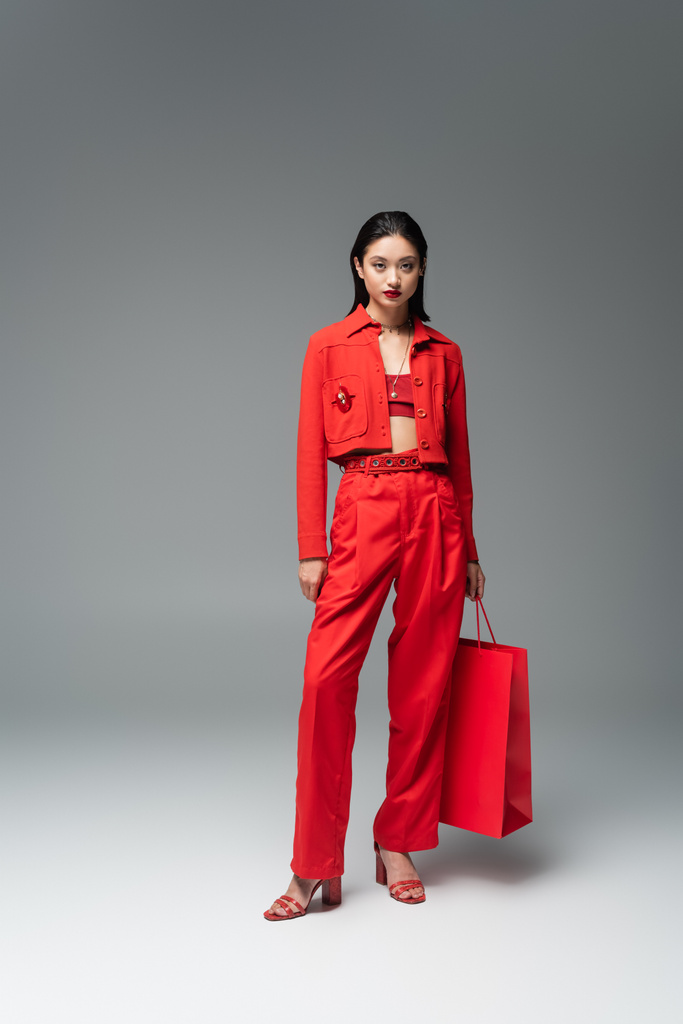 повна довжина азіатської моделі в червоній куртці і штанах, що стоять з сумкою для покупок на сірому фоні
 - Фото, зображення