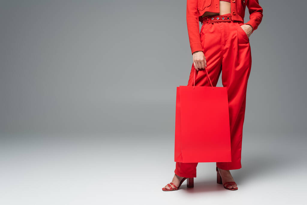 cropped view of woman holding shopping bag while standing with hand in pocket of red pants on grey background - Photo, Image