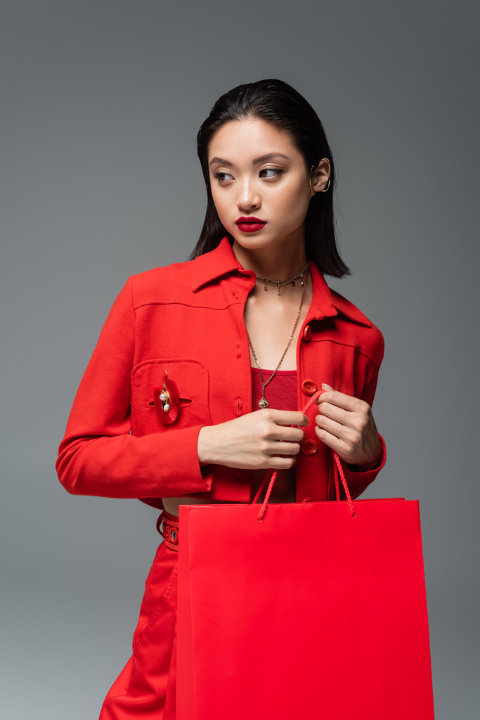 Asiatin in roter Jacke und Halsketten hält Einkaufstasche in der Hand und schaut isoliert auf grau - Foto, Bild