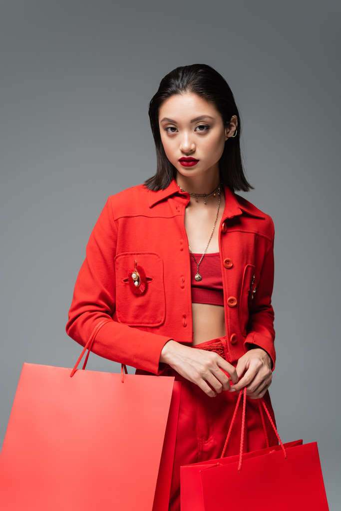 modna azjatycka kobieta w czerwonej kurtce trzymając torby na zakupy i patrząc na aparat odizolowany na szary - Zdjęcie, obraz