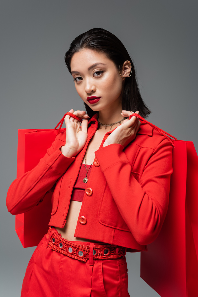 mulher asiática bonita na jaqueta da moda segurando sacos de compras vermelhos e olhando para a câmera isolada no cinza - Foto, Imagem
