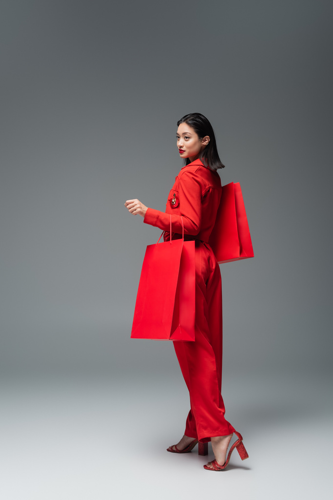 full length of fashionable asian woman with red shopping bags looking away on grey background - Photo, Image