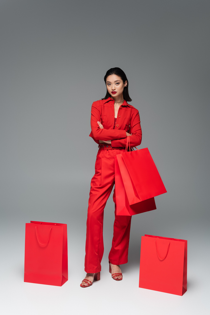 πλήρες μήκος της ασιατικής γυναίκας στο κόκκινο κομψό κοστούμι στέκεται με σταυρωμένα χέρια κοντά τσάντες ψώνια σε γκρι φόντο - Φωτογραφία, εικόνα