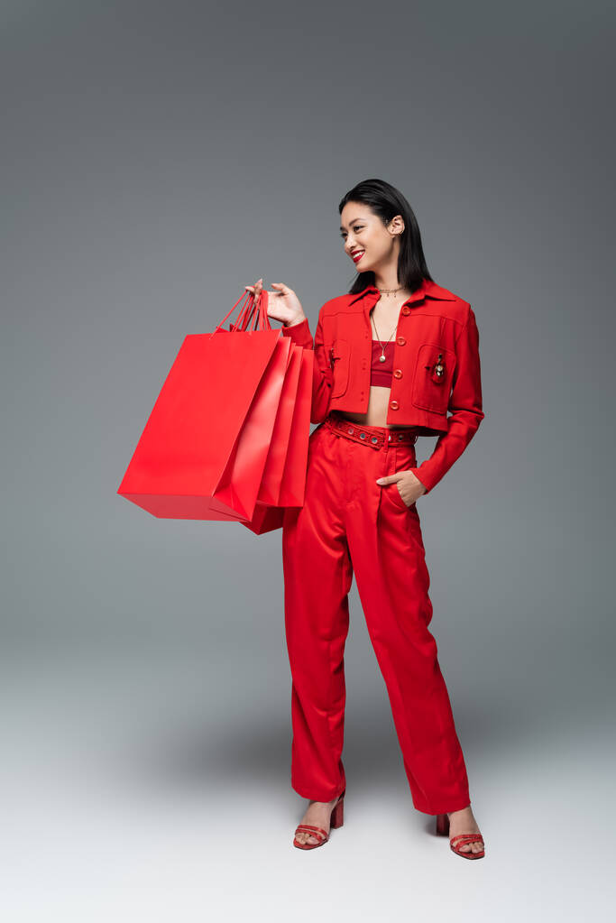 täyspitkä aasialainen nainen punainen tyylikäs asu poseeraa ostoskassit ja käsi taskussa harmaalla taustalla - Valokuva, kuva