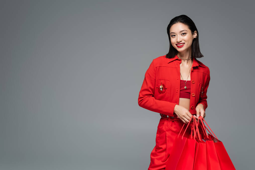 muodikas aasialainen nainen hymyilee kameralle seisoessaan punaiset ostoskassit eristetty harmaa - Valokuva, kuva