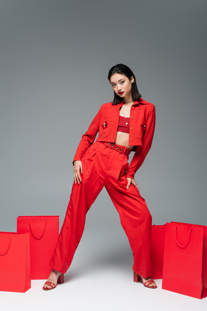 teljes hossza ázsiai modell piros kabát és nadrág pózol közelében bevásárló táskák szürke háttér - Fotó, kép