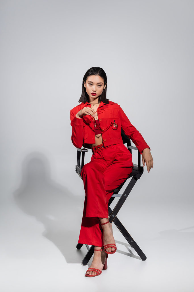 полная длина азиатской модели в красной куртке и брюках, держащих стильные солнцезащитные очки, сидя на стуле на сером фоне - Фото, изображение