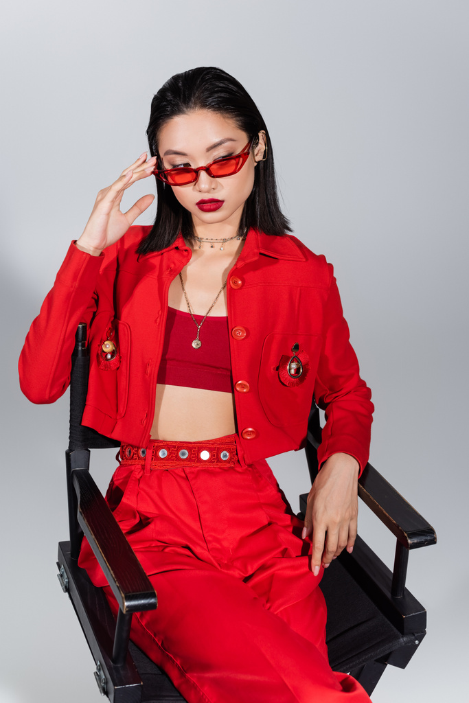 модна азіатська жінка в червоному вбранні регулює сонцезахисні окуляри, сидячи на стільці ізольовано на сірому
 - Фото, зображення