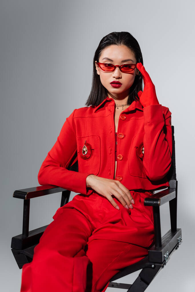 modelo asiático em terno vermelho tocando óculos de sol elegantes enquanto sentado isolado no cinza - Foto, Imagem