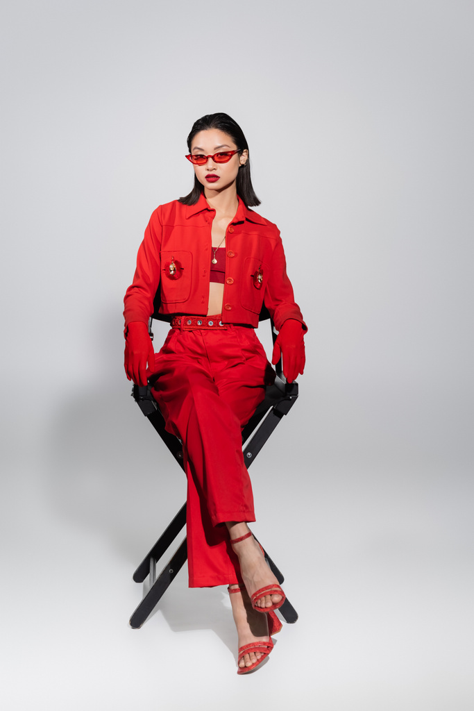 teljes hossza ázsiai nő piros elegáns napszemüveg és elegáns öltöny ül széken szürke háttér - Fotó, kép