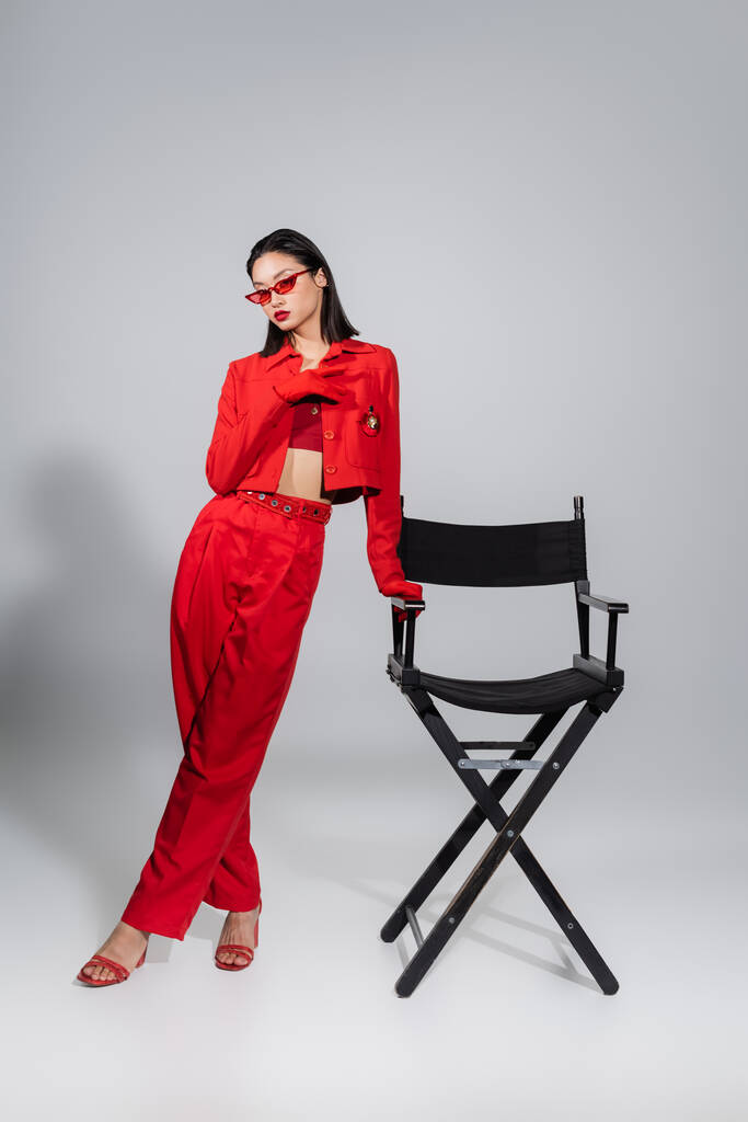 full length of brunette asian woman in red suit and sunglasses posing near chair on grey background - Photo, Image