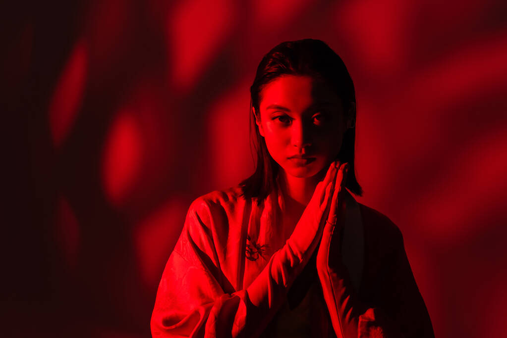 Kimono pelerinli genç Asyalı kadın dua eden elleri kırmızı ışıkta, koyu arka planda duruyor. - Fotoğraf, Görsel