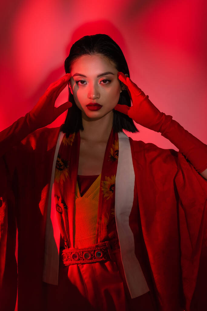 mladá asijská žena v kimono pláštěnku a rukavice pózování s rukama blízko obličeje na abstraktním pozadí s červeným světlem - Fotografie, Obrázek