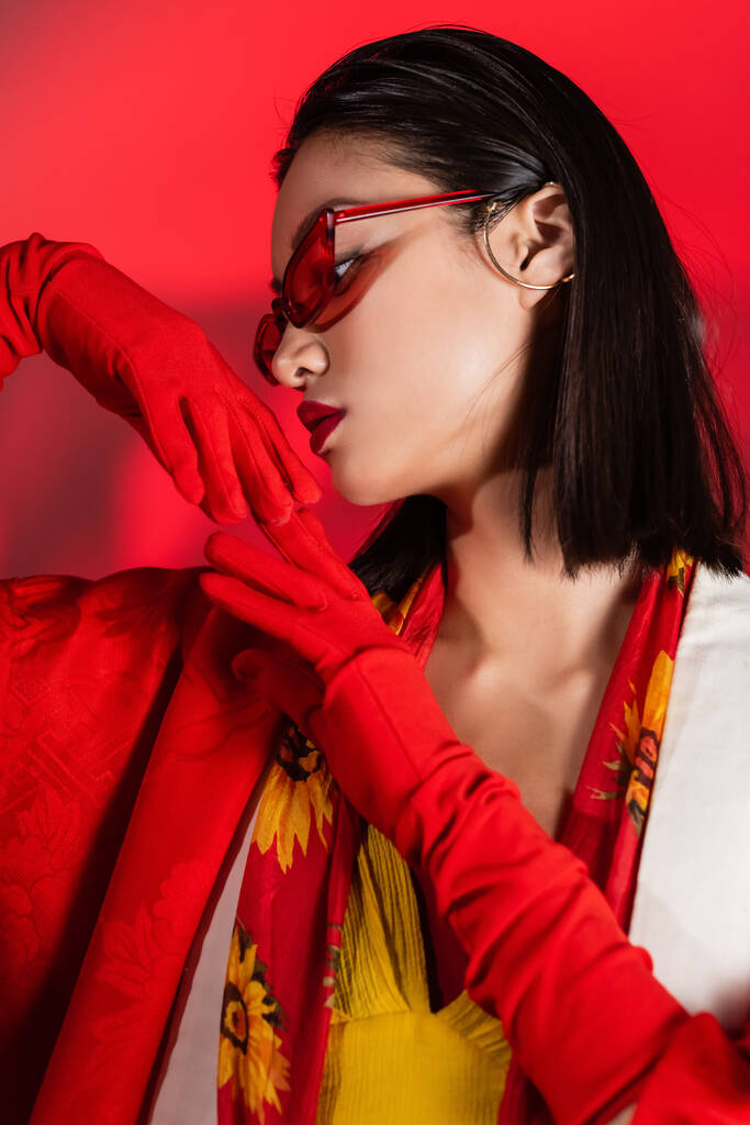 érzéki ázsiai nő napszemüvegben és nyakkendő virágmintás pózol kesztyűben piros háttér - Fotó, kép