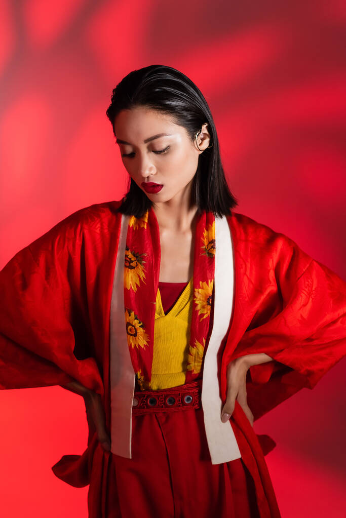 brunette asiatique femme en foulard avec décor floral et kimono cape posant avec les mains sur les hanches sur fond rouge abstrait - Photo, image