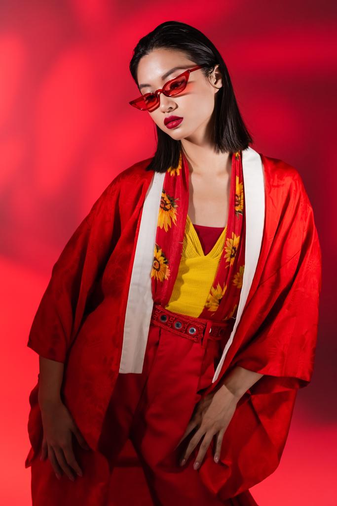 divatos ázsiai nő kimonó köpenyben és napszemüveg pózol a piros háttér árnyék - Fotó, kép