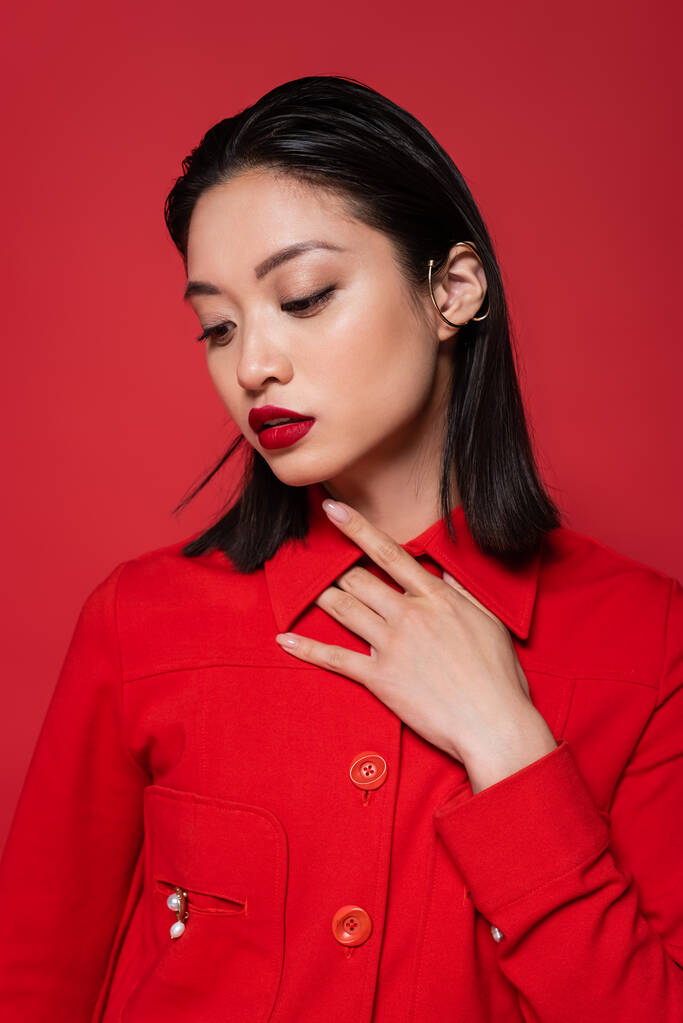 sensual asian woman in trendy jacket and ear cuff posing with hand on chest isolated on red - Photo, Image