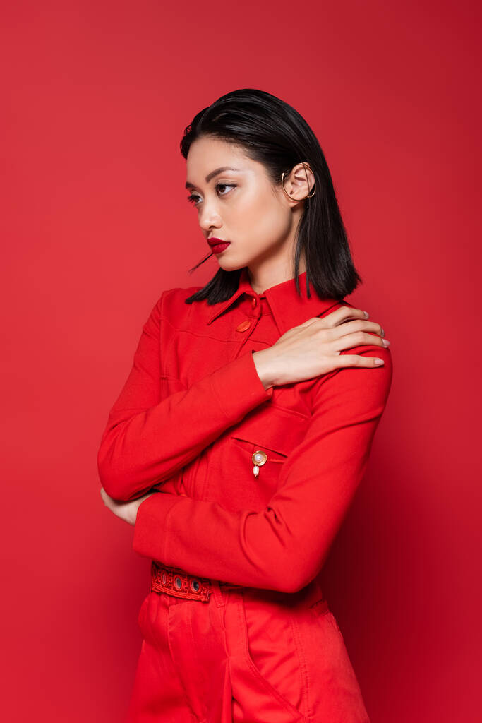 μοντέρνα Ασιάτισσα με σακάκι που ποζάρει με το χέρι στον ώμο και κοιτάζει μακριά απομονωμένη στο κόκκινο - Φωτογραφία, εικόνα