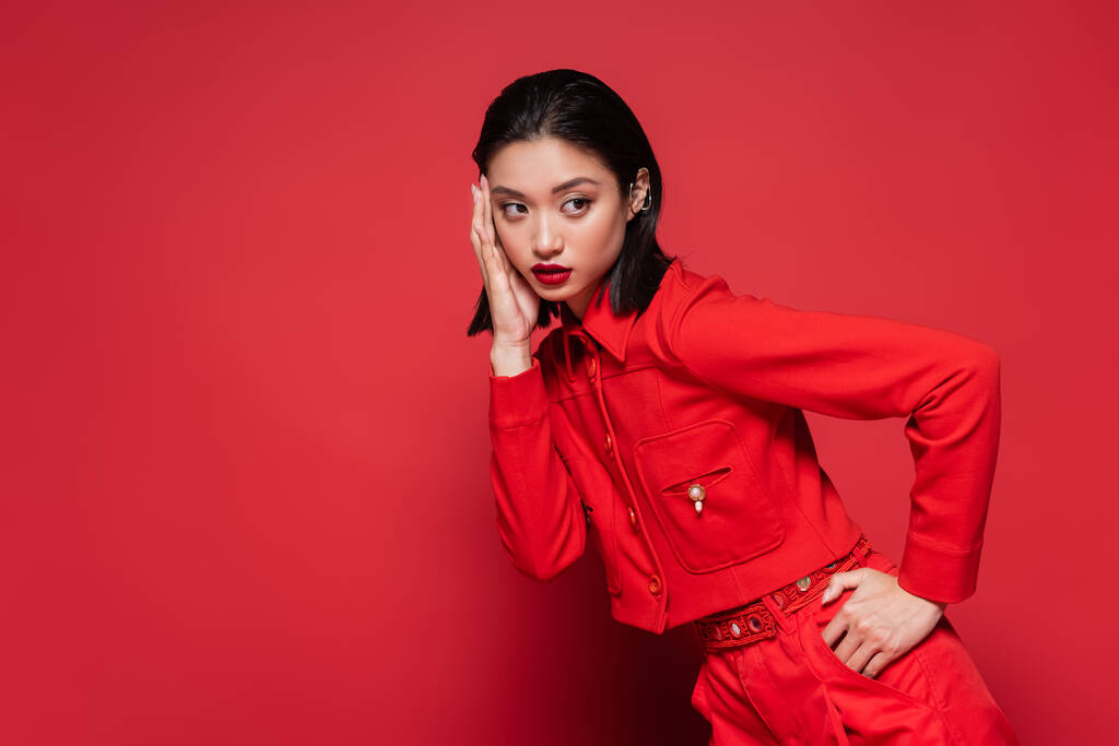 young asian model in elegant attire and ear cuff touching face and holding hand on hip on red background - Photo, Image