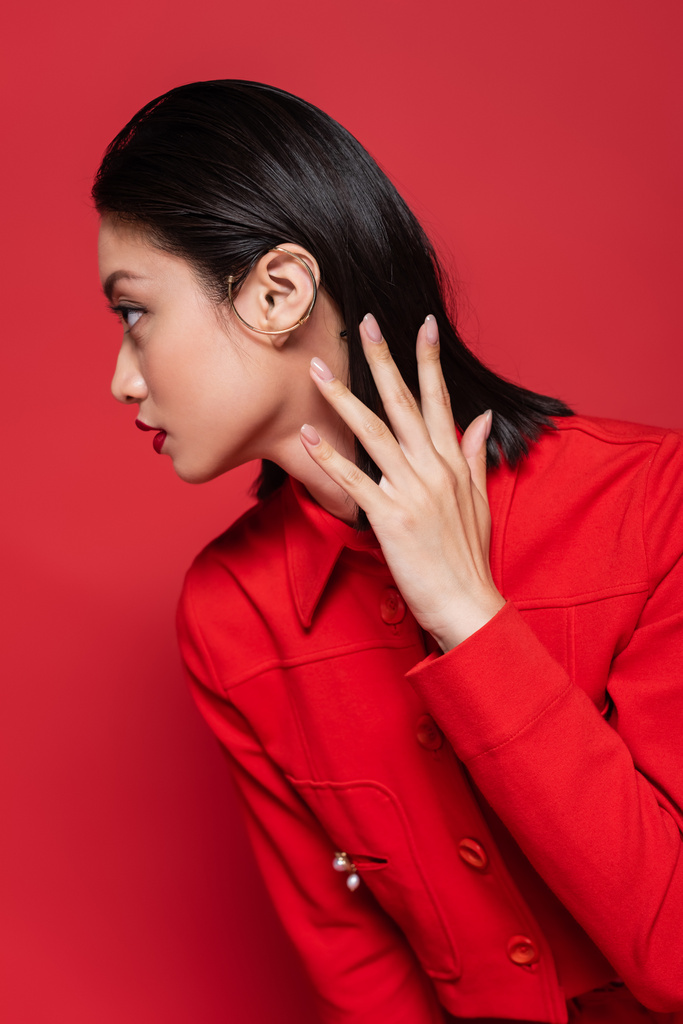 elegante asiatische Frau in Jacke und Ohrenmanschette, die die Hand am Hals hält und isoliert auf Rot wegschaut - Foto, Bild