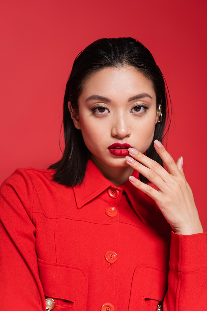 portret van mooie aziatische vrouw in stijlvolle jas kijken naar camera en aanraken lippen geïsoleerd op rood - Foto, afbeelding