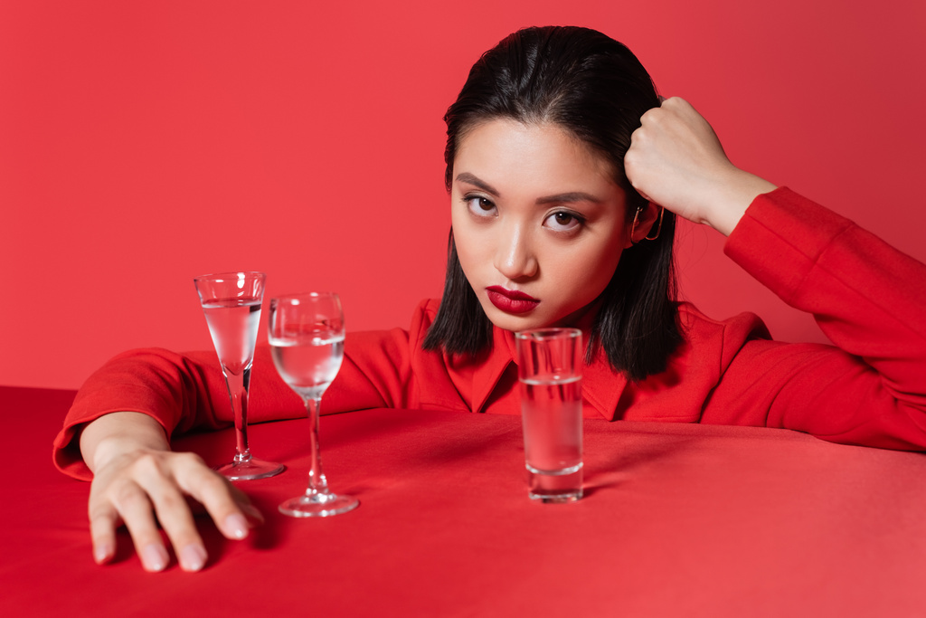 young asian woman in jacket holding hand near head and looking at camera near glasses with water isolated on red - Photo, Image