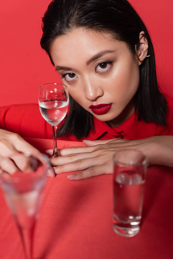 bruna donna asiatica con trucco e bracciale per le orecchie guardando la fotocamera vicino agli occhiali con acqua in primo piano offuscata isolata sul rosso - Foto, immagini