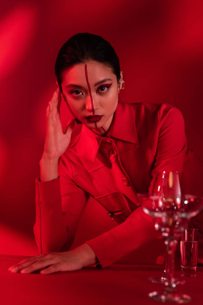 азиатка с творческим обликом, позирующая в модной куртке рядом с размытыми очками с водой на красном затененном фоне - Фото, изображение