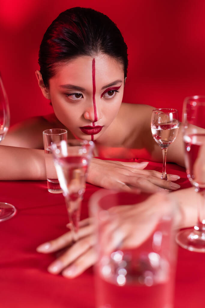sinnliche asiatische Frau mit kreativem Gesicht und nackten Schultern, die in der Nähe von Wassergläsern auf rotem Hintergrund posiert - Foto, Bild