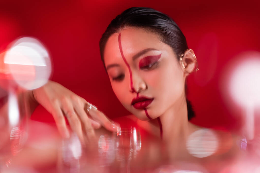 morena mujer asiática con rostro artístico cerca de gafas borrosas sobre fondo rojo - Foto, imagen
