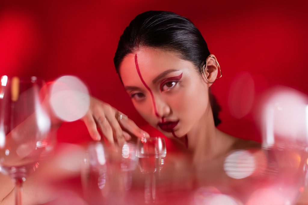 giovane donna asiatica con trucco creativo guardando la fotocamera vicino a occhiali sfocati su sfondo rosso - Foto, immagini